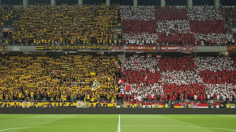 Packed football stadium at night, one end filled with yellow and black supporters and the other with red and white, a ball on the centre spot